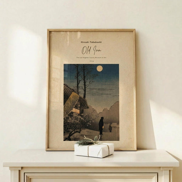 May include: A framed vintage Japanese woodblock print showing a moonlit evening scene with a traditional thatched-roof inn, bare tree, and silhouetted figures on a path. The artwork features muted blues, browns, and warm golden light from a window. Natural wood frame with cream matting, displayed on a light wooden surface alongside a small white wrapped gift with twine and dried botanicals.