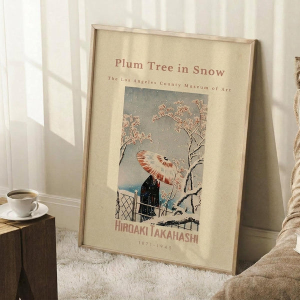 May include: Framed art print leaning against white wall on white textured rug, showing Japanese woodblock style winter scene with figure holding umbrella under plum tree branches in snow. Cream-colored matting with museum attribution text. Small wooden block with white coffee cup nearby. Natural lighting creates soft shadows.