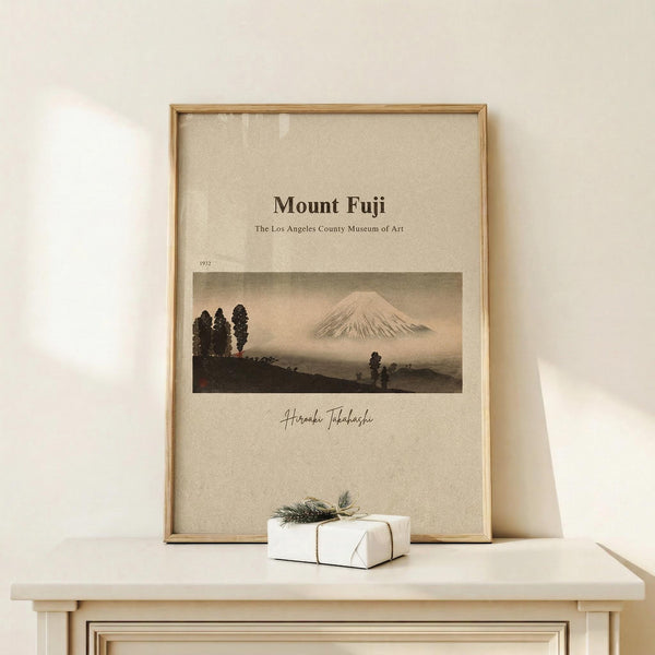 May include: Framed vintage-style poster featuring Mount Fuji landscape with trees in foreground, displayed on light wood shelf against cream wall. Natural wood frame surrounds beige poster with Japanese mountain scene in muted earth tones. Small white wrapped gift with twine and pine sprig sits beside frame.