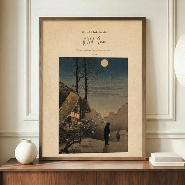 May include: Framed vintage Japanese woodblock print showing a moonlit evening scene with traditional thatched-roof buildings, bare trees, and silhouetted figures on a path. The artwork features muted blues, browns, and cream tones with a full moon in the sky. Displayed on a wooden sideboard with white ceramic vase and books, against cream walls with white molding.