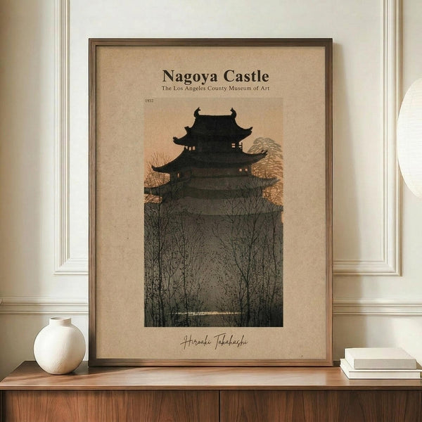 May include: Framed vintage-style poster featuring a traditional Japanese castle silhouette against a warm beige background, displayed on a wooden credenza alongside a white ceramic vase and stacked books. The artwork shows a multi-tiered pagoda structure emerging from bare winter trees, rendered in dark tones with subtle orange accents. The piece is matted in a brown wooden frame and positioned against white wainscoting in an elegant interior setting.