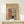 May include: Framed vintage Japanese woodblock print poster showing a snowy winter scene with red torii gate, traditional buildings, and two figures in kimono. Print has beige background with muted colors including red, blue, and cream tones. Displayed in thin wooden frame on light wood surface next to small wrapped white gift box against cream wall.
