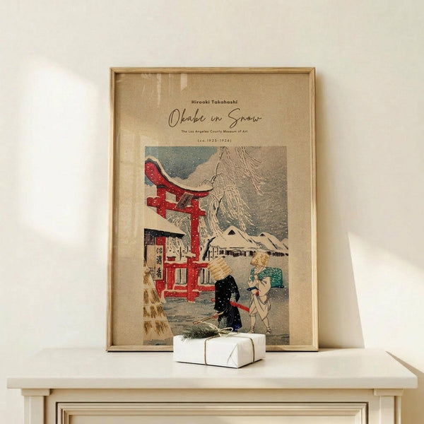 May include: Framed vintage Japanese woodblock print poster showing a snowy winter scene with red torii gate, traditional buildings, and two figures in kimono. Print has beige background with muted colors including red, blue, and cream tones. Displayed in thin wooden frame on light wood surface next to small wrapped white gift box against cream wall.