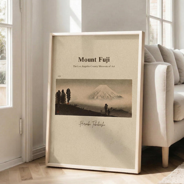 May include: Framed vintage-style poster featuring Mount Fuji landscape in muted earth tones, displayed in bright corner room with beige armchair, white walls, and natural lighting from windows. The artwork shows the iconic mountain with trees in foreground against cream background, housed in light wood frame leaning against wall on hardwood floors.