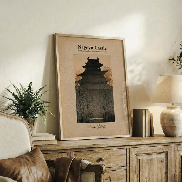 May include: Framed vintage-style poster featuring a dark silhouette of a traditional Japanese castle surrounded by bare winter trees against a warm beige background. The artwork is displayed on a rustic wooden sideboard alongside a fern plant, ceramic vase, table lamp, and books in a cozy living room setting with neutral tones and natural textures.