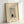 May include: Framed vintage Japanese woodblock print poster featuring a figure with parasol in snowy winter scene with flowering plum tree branches. Natural wood frame against white wall corner, showing muted colors of gray, pink, and blue with delicate snowfall details and traditional artistic style.
