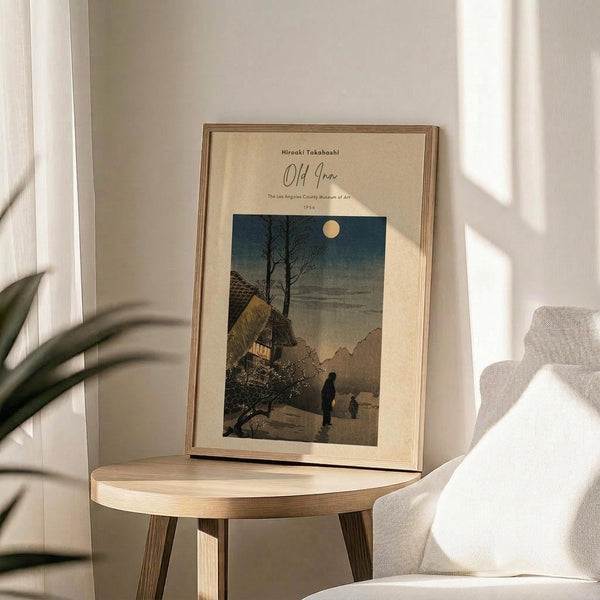 May include: Japanese woodblock print poster in wooden frame showing moonlit village scene with traditional houses, bare tree, and two figures walking on snowy path under full moon. Displayed on round wooden side table next to beige upholstered chair with decorative pillow. Soft natural lighting creates shadows across white wall. Plant leaves visible in corner. Vintage-style artwork features blue night sky transitioning to warm earth tones below.