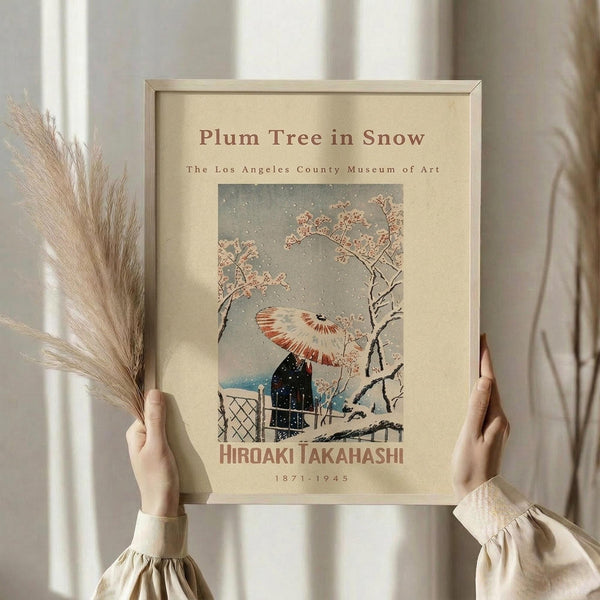 May include: Hands holding a framed vintage Japanese woodblock print showing a snow-covered plum tree with pink blossoms, traditional umbrella, and small building during snowfall. The artwork features muted colors on cream background within a light wood frame. Dried pampas grass visible to the left against white curtained window backdrop.