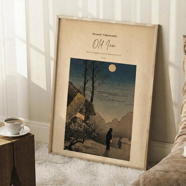May include: Framed vintage Japanese woodblock print showing a moonlit mountain village scene with traditional thatched roof buildings, bare trees, and silhouetted figures on a path. The artwork features muted blues, browns, and cream tones in a beige mat and light wood frame. Displayed leaning against white paneled wall on textured white rug, with wooden side table holding white coffee cup and saucer, and beige knit throw blanket visible.