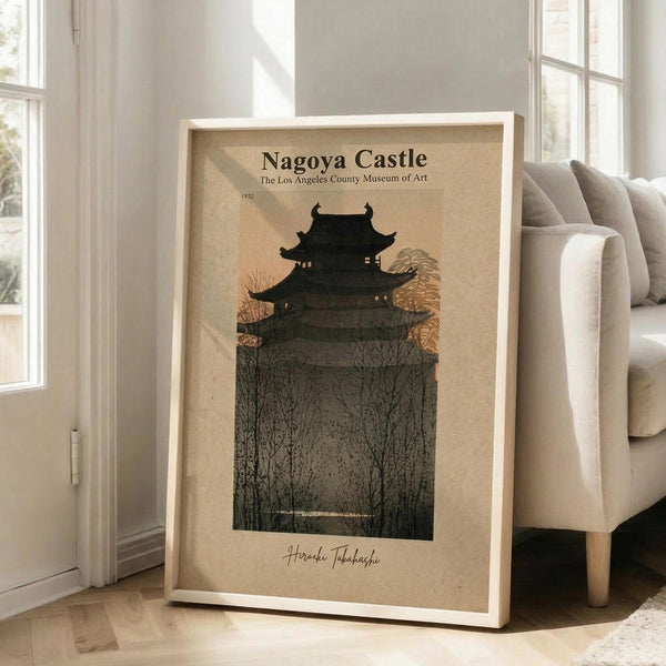 May include: Framed vintage-style art print featuring a traditional multi-tiered Japanese castle silhouetted against autumn trees with golden foliage. The poster has a cream background with dark green and brown tones, displayed in a light wood frame leaning against a white wall next to a beige upholstered chair in a bright, minimalist room with hardwood floors.