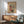 May include: Japanese woodblock print poster in wooden frame hanging above modern credenza, showing snowy winter scene with red torii gate, traditional buildings, and figures with umbrellas. Mid-century mint green armchair with wooden arms positioned nearby, white table lamp on sideboard, natural sunlight casting shadows across white wall in contemporary living room setting.