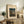 May include: Framed vintage Japanese woodblock print showing a moonlit evening scene with traditional thatched-roof house, bare tree, and silhouetted figures on a path. Print displays in cream matting with light wood frame, styled on rustic wooden sideboard alongside fern plant, ceramic vase, stacked books, and beige upholstered chair in warm, neutral-toned living room setting.