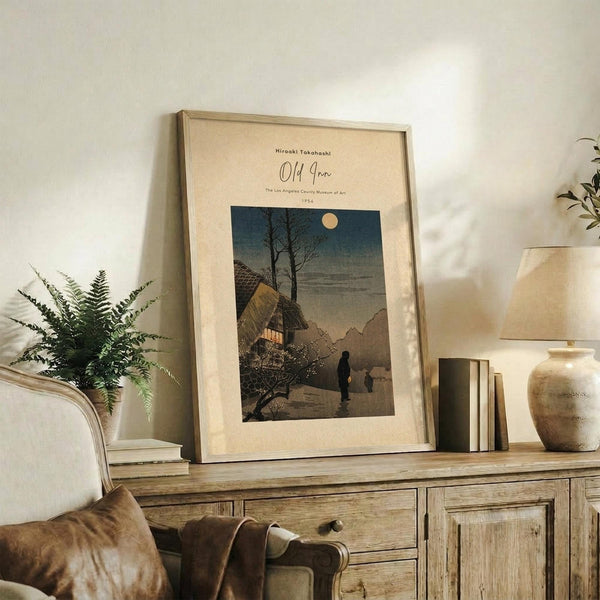 May include: Framed vintage Japanese woodblock print showing a moonlit evening scene with traditional thatched-roof house, bare tree, and silhouetted figures on a path. Print displays in cream matting with light wood frame, styled on rustic wooden sideboard alongside fern plant, ceramic vase, stacked books, and beige upholstered chair in warm, neutral-toned living room setting.