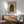 May include: Framed vintage-style poster of Japanese castle silhouette against twilight sky with bare winter trees, displayed above light wood sideboard with white ceramic lamp in modern living room setting. Mid-century sage green upholstered chair with wooden arms visible in foreground. Natural sunlight creates dramatic shadows across white wall and warm wood furniture.