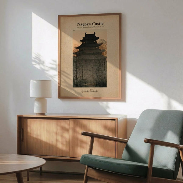May include: Framed vintage-style poster of Japanese castle silhouette against twilight sky with bare winter trees, displayed above light wood sideboard with white ceramic lamp in modern living room setting. Mid-century sage green upholstered chair with wooden arms visible in foreground. Natural sunlight creates dramatic shadows across white wall and warm wood furniture.