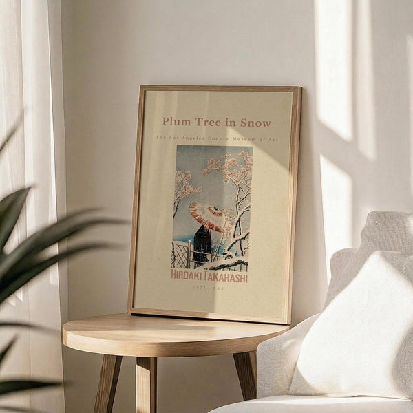 May include: Framed vintage Japanese art print featuring a figure with red and white umbrella walking beneath snow-covered plum tree branches. Cream-colored poster with museum exhibition text displayed on round wooden side table next to beige upholstered chair. Soft natural lighting creates shadows across white wall, with green plant leaves visible in corner.