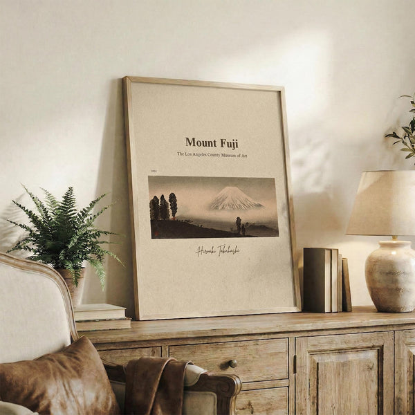 May include: Framed vintage-style art print featuring Mount Fuji with silhouetted trees in muted earth tones, displayed on rustic wooden sideboard alongside fern plant, stacked books, and ceramic vase in neutral-toned living room setting with beige chair and pillow visible