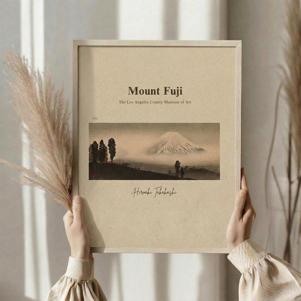 May include: Hands holding a framed vintage Japanese art print showing Mount Fuji with snow-capped peak rising above dark silhouetted trees and rolling hills in muted earth tones. The beige matted artwork is displayed in a light wood frame against soft natural lighting with dried pampas grass visible to the left side.
