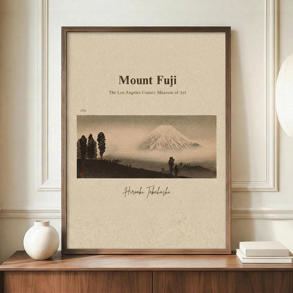 May include: Framed vintage-style poster featuring Mount Fuji with snow-capped peak emerging from misty clouds, dark silhouetted trees in foreground, rendered in muted earth tones. Museum exhibition format with beige background, date 1932, and artist signature. Displayed on wooden console alongside white ceramic vase and stacked books against cream wall with white trim molding.