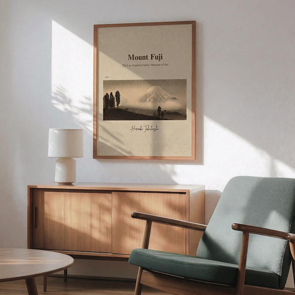 a framed poster of Mount Fuji hanging on a wall, with a wooden cabinet below it and a green armchair in the foreground.