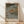 May include: Framed Japanese woodblock print featuring autumn landscape with orange and green trees along a river, small figures on wooden dock, traditional ukiyo-e style artwork in natural wood frame, displayed against white wall on light wooden floor with soft natural lighting creating diagonal shadows