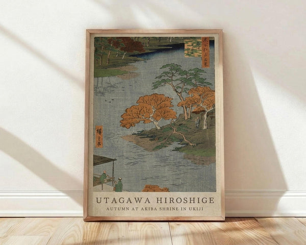 May include: Framed Japanese woodblock print featuring autumn landscape with orange and green trees along a river, small figures on wooden dock, traditional ukiyo-e style artwork in natural wood frame, displayed against white wall on light wooden floor with soft natural lighting creating diagonal shadows