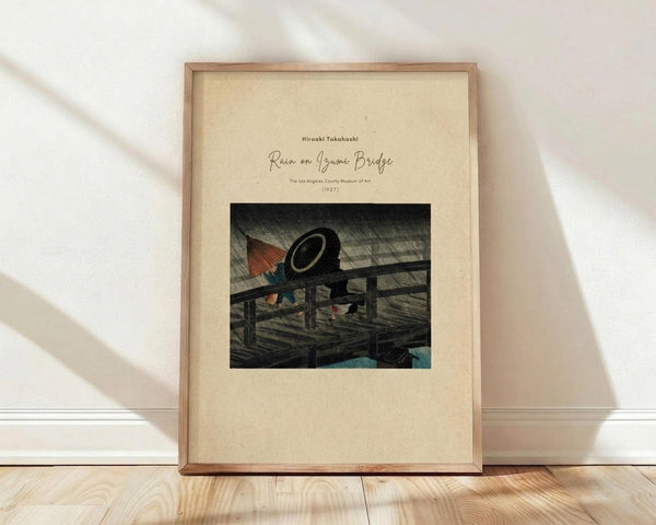 May include: Framed art print displaying a Japanese woodblock scene of figures with umbrellas on a wooden bridge during rainfall, rendered in muted greens and earth tones. The cream-colored mat features elegant script text above the artwork. The thin wooden frame rests against a white wall on light hardwood flooring, with soft natural lighting creating gentle shadows across the scene.