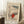 May include: Japanese woodblock print poster in light wood frame leaning against white wall corner, showing figures in red winter clothing walking through snowy mountain landscape with bare trees and falling snow. Small wooden side table with white coffee cup nearby on textured cream rug, cozy beige blanket visible at edge.
