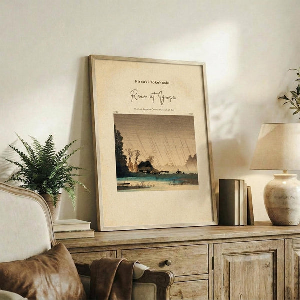 May include: Framed vintage Japanese woodblock print showing a rainy landscape with traditional house, bare trees, and diagonal rain lines against muted earth tones. Print displayed on rustic wooden sideboard alongside fern plant, ceramic vase, and books in warm, neutral-toned living room setting.