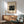 May include: Framed vintage Japanese woodblock print showing a wooden bridge in rain with figures holding umbrellas, displayed above a mid-century modern wood credenza with white ceramic lamp. Light green upholstered armchair with wooden frame sits nearby. Warm natural lighting creates shadows across white wall in minimalist room setting.