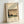 May include: Framed vintage Japanese woodblock print showing a rainy landscape scene with traditional house, bare trees, and turquoise water under diagonal rain lines across sepia sky. Light wood frame sits propped against white wall on light surface, creating soft shadows.