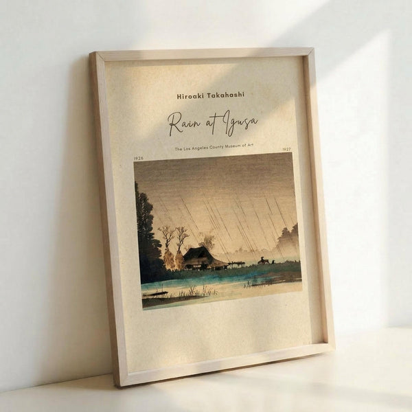 May include: Framed vintage Japanese woodblock print showing a rainy landscape scene with traditional house, bare trees, and turquoise water under diagonal rain lines across sepia sky. Light wood frame sits propped against white wall on light surface, creating soft shadows.