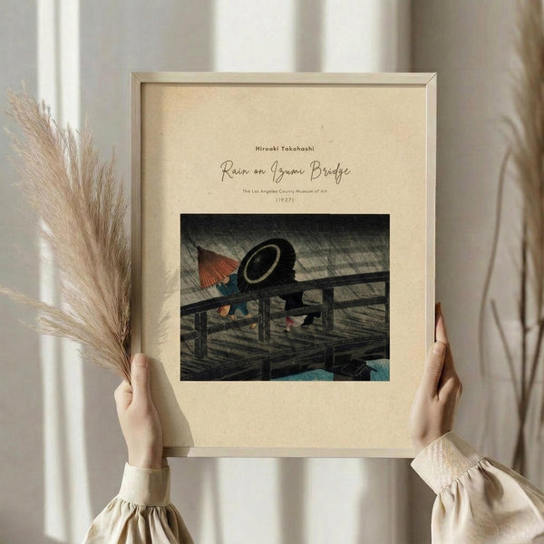 May include: Person in cream sweater holding framed Japanese woodblock print showing figures with umbrellas crossing wooden bridge in rain, displayed against white curtained window with dried pampas grass stems, neutral beige mat and light wood frame
