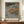May include: Japanese woodblock print poster in wooden frame displayed on walnut credenza beside white ceramic vase and stacked books. Artwork depicts autumn landscape with orange-leafed trees along water, small figures in boat, traditional red text cartouches, and "UTAGAWA HIROSHIGE AUTUMN AT AKIBA SHRINE IN UKIJI" caption below image.