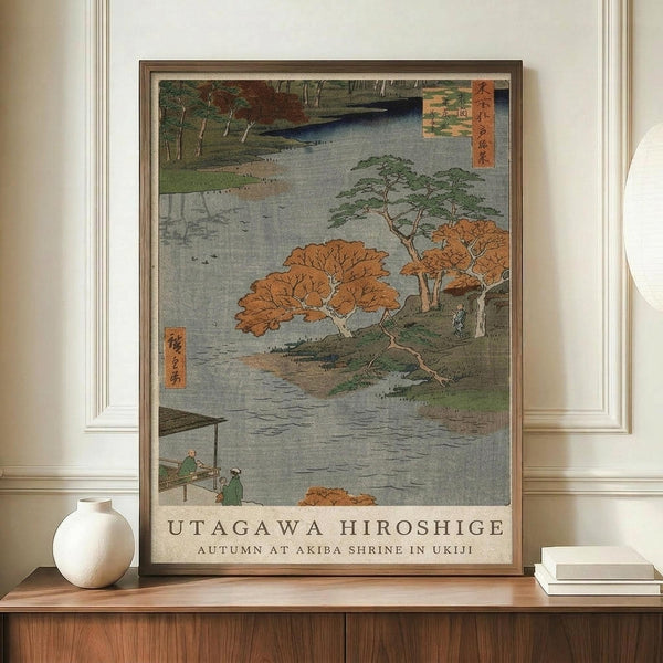 May include: Japanese woodblock print poster in wooden frame displayed on walnut credenza beside white ceramic vase and stacked books. Artwork depicts autumn landscape with orange-leafed trees along water, small figures in boat, traditional red text cartouches, and "UTAGAWA HIROSHIGE AUTUMN AT AKIBA SHRINE IN UKIJI" caption below image.
