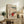 May include: Framed vintage Japanese woodblock print showing a figure in red robes walking through falling snow on a mountain path, displayed on rustic wooden sideboard alongside fern plant, ceramic vase, and stacked books in neutral-toned living room setting with cream chair visible.