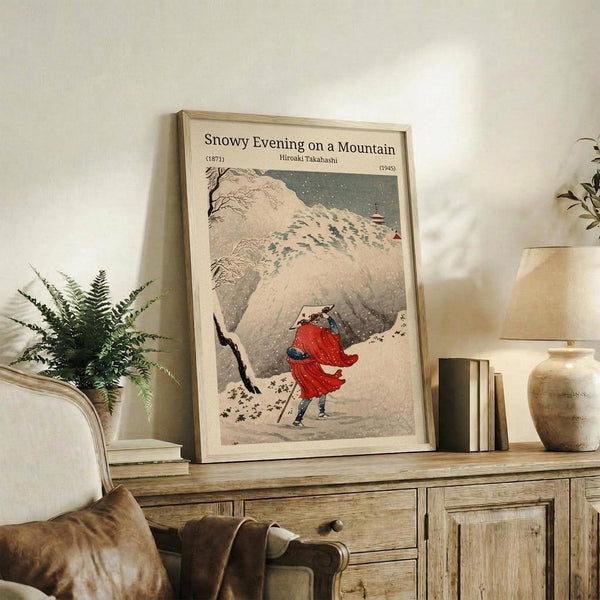 May include: Framed vintage Japanese woodblock print showing a figure in red robes walking through falling snow on a mountain path, displayed on rustic wooden sideboard alongside fern plant, ceramic vase, and stacked books in neutral-toned living room setting with cream chair visible.