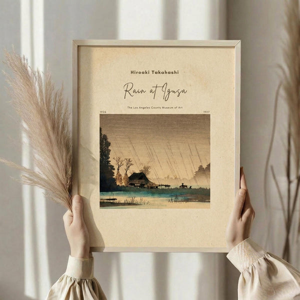 May include: Hands holding a framed vintage Japanese woodblock print showing a rainy landscape with traditional buildings, trees, and water in muted earth tones. The artwork is displayed in a light wooden frame against cream matting. Person wears a beige ruffled sleeve top and holds dried pampas grass, with soft natural lighting and white curtains in the background.