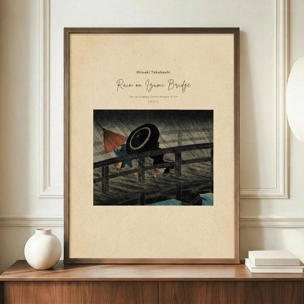 May include: Framed vintage Japanese woodblock print showing a wooden bridge in rain with colorful umbrellas and a large black hat, displayed on wooden sideboard beside white ceramic vase and stacked books against cream paneled wall in modern interior setting.