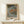 May include: Japanese woodblock print poster in light wooden frame showing autumn riverside scene with orange and green trees, small figures by water, and Japanese text. Displayed on pale wooden surface against cream wall with small white wrapped gift beside frame.