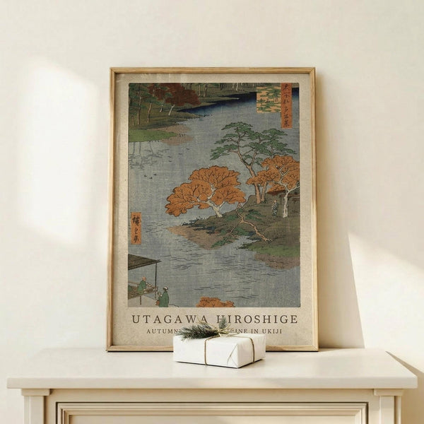 May include: Japanese woodblock print poster in light wooden frame showing autumn riverside scene with orange and green trees, small figures by water, and Japanese text. Displayed on pale wooden surface against cream wall with small white wrapped gift beside frame.
