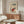 May include: Japanese woodblock print poster displayed above wooden sideboard in modern living room. Artwork shows figures in red clothing walking through snowy mountain landscape with falling snow. White upholstered chair and glass coffee table visible in foreground, with decorative lamp on sideboard. Print appears framed in thin brown frame against cream colored wall.