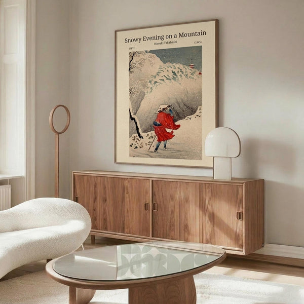 May include: Japanese woodblock print poster displayed above wooden sideboard in modern living room. Artwork shows figures in red clothing walking through snowy mountain landscape with falling snow. White upholstered chair and glass coffee table visible in foreground, with decorative lamp on sideboard. Print appears framed in thin brown frame against cream colored wall.