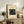 May include: Framed beige poster with Japanese woodblock print showing figures with umbrellas on a wooden bridge in rain, displayed on rustic wooden sideboard alongside fern plant, ceramic vase, and stacked books in neutral-toned living room setting with upholstered chair visible.