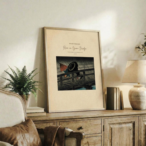 May include: Framed beige poster with Japanese woodblock print showing figures with umbrellas on a wooden bridge in rain, displayed on rustic wooden sideboard alongside fern plant, ceramic vase, and stacked books in neutral-toned living room setting with upholstered chair visible.