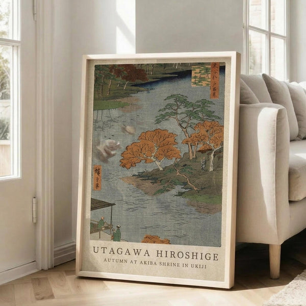 May include: Japanese woodblock print poster in light wooden frame showing autumn landscape with orange and red foliage trees beside a river, small figures on a boat, traditional Japanese characters in corner, displayed on wooden floor against white wall next to beige upholstered chair in bright room with large windows.