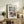 May include: Framed vintage Japanese woodblock print showing a snow-covered temple gate with traditional red and white architecture surrounded by winter trees. The beige-matted artwork sits on a rustic wooden sideboard alongside books, a ceramic vase, and fern plant. Warm lighting creates a cozy living room atmosphere with neutral tones throughout the styled scene.