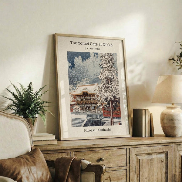 May include: Framed vintage Japanese woodblock print showing a snow-covered temple gate with traditional red and white architecture surrounded by winter trees. The beige-matted artwork sits on a rustic wooden sideboard alongside books, a ceramic vase, and fern plant. Warm lighting creates a cozy living room atmosphere with neutral tones throughout the styled scene.