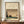 May include: Framed vintage Japanese woodblock print reproduction showing a rainy landscape scene with diagonal rain lines, traditional buildings near water, trees, and small figures. The artwork has muted earth tones in brown, green, and beige. Displayed on a wooden sideboard next to a white ceramic vase and stack of books against cream walls with white trim molding.