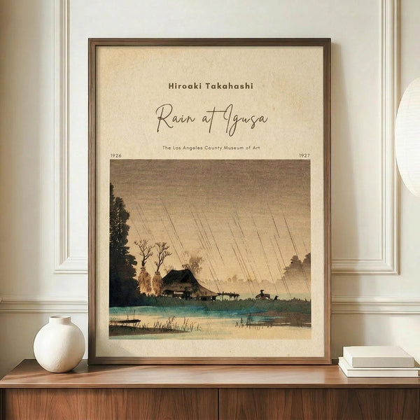 May include: Framed vintage Japanese woodblock print reproduction showing a rainy landscape scene with diagonal rain lines, traditional buildings near water, trees, and small figures. The artwork has muted earth tones in brown, green, and beige. Displayed on a wooden sideboard next to a white ceramic vase and stack of books against cream walls with white trim molding.